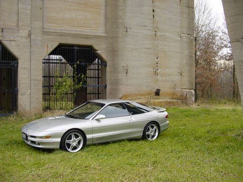 1994 Ford Probe - A silver 1994 Ford Probe is parked on a grassy area next to a large concrete structure.