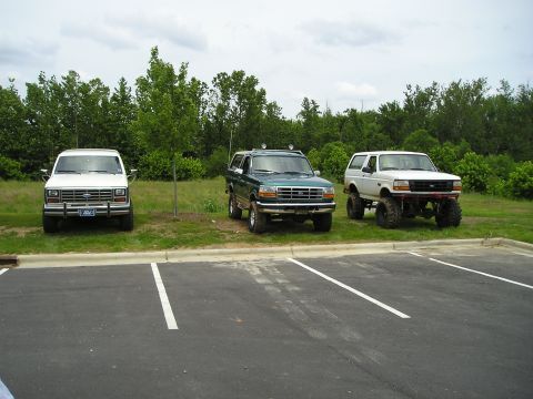 1996 Ford Bronco - Three Ford Broncos are parked in a lot with trees and grass in the background.