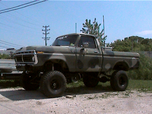 Camo-painted lifted 1970s Ford pickup on big off-road tires, gravel lot.