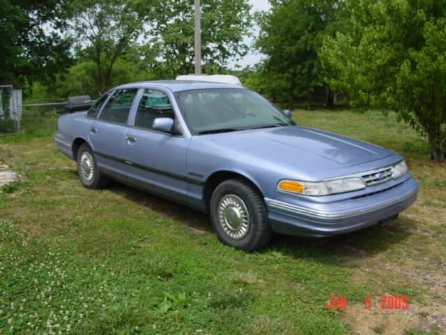 Light blue Ford Crown Victoria sedan parked on grass, front three-quarter passenger view