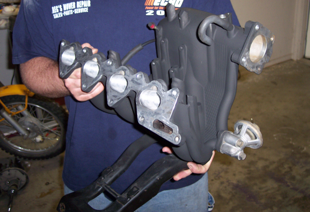Person holding black plastic intake manifold with aluminum flange in a garage