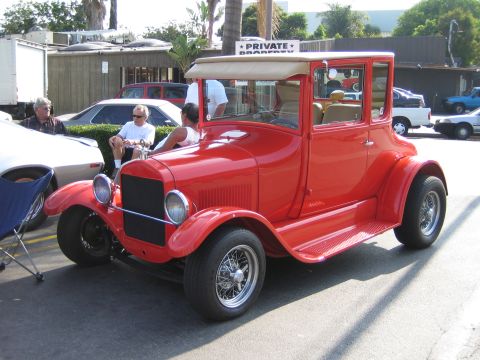 1985 Ford Mustang - A vintage red coupe is parked outdoors, with people sitting nearby and other vehicles in the background.