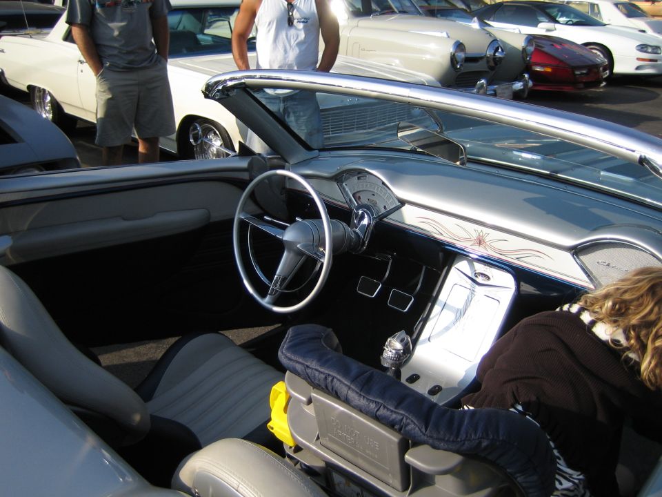 Interior view of a classic convertible showing dashboard, steering wheel, and seats. Other cars are visible in the background
