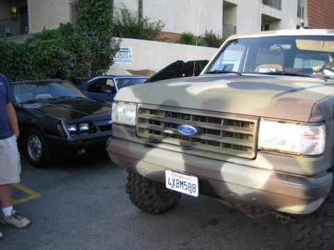 1985 Ford Mustang - A camouflage-painted Ford Bronco is parked in front of a black convertible.