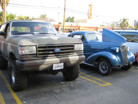 1985 Ford Mustang - A camouflaged Ford Bronco is parked next to a blue classic car. The Bronco's front end and large tires are visible.