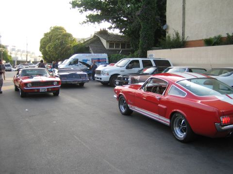 1985 Ford Mustang - A red classic coupe with white racing stripes is parked on a street alongside other vehicles.