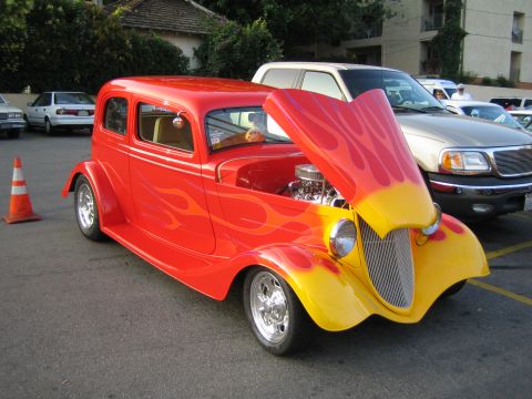 1985 Ford Mustang - A custom hot rod sedan with flame paint and an open hood is parked in a lot with other vehicles.