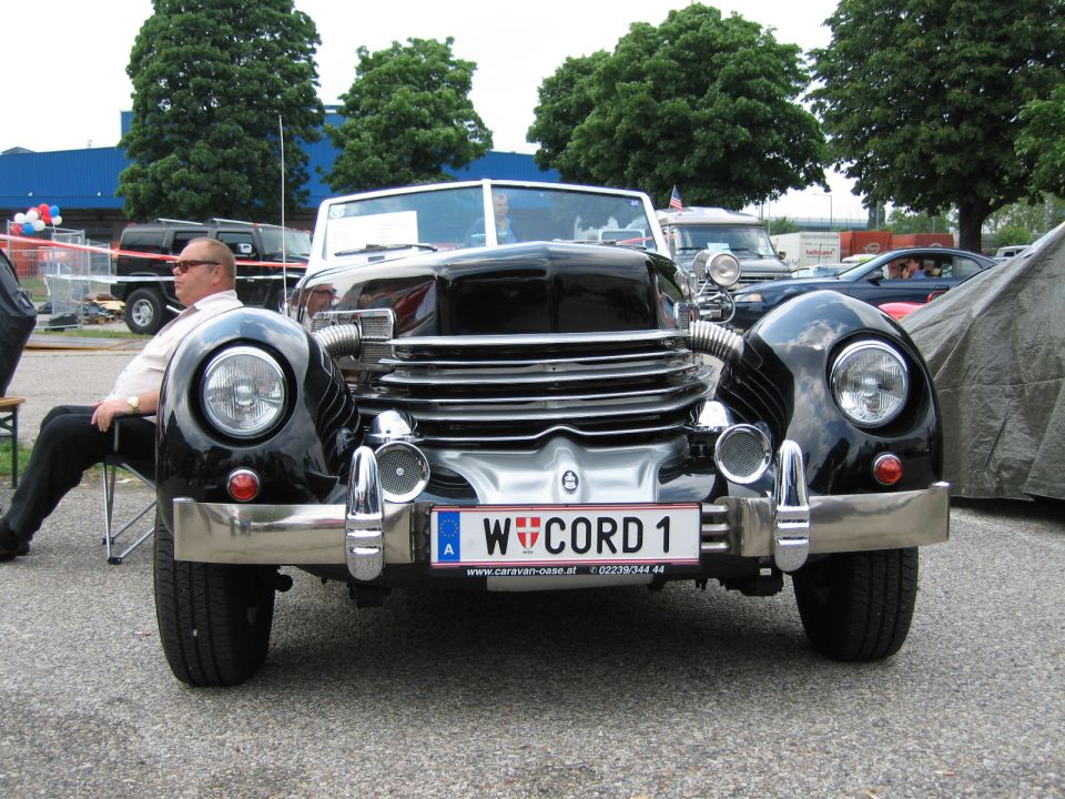 Front view of a black Cord 810/812 convertible with a man sitting in a chair to the left