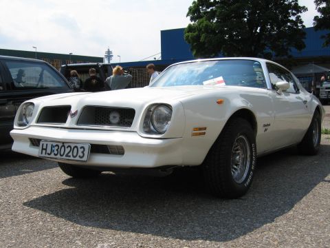 1992 Ford Crown Victoria - A white 1977 Pontiac Firebird coupe is parked outdoors, with other vehicles and people in the background.