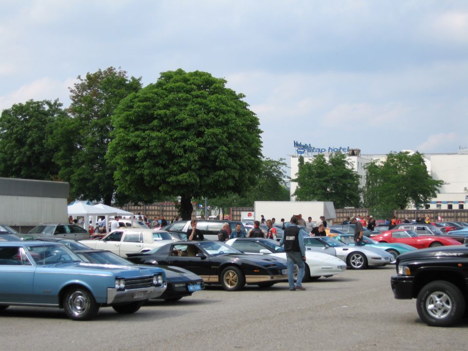 Outdoor car show scene with various vehicles parked, including a blue classic car and a black pickup truck in the foreground