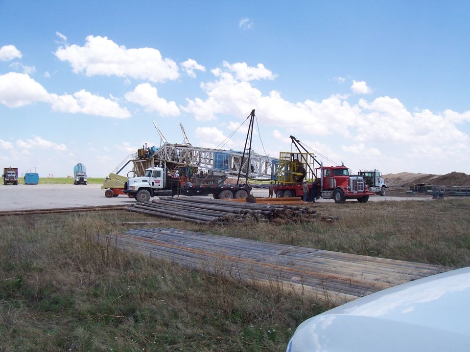 Oilfield workover site with semi-trucks, drilling rig mast and stacked steel pipe under blue sky