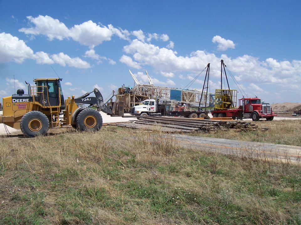 Yellow John Deere loader and red truck at a construction site with a large rig in the background