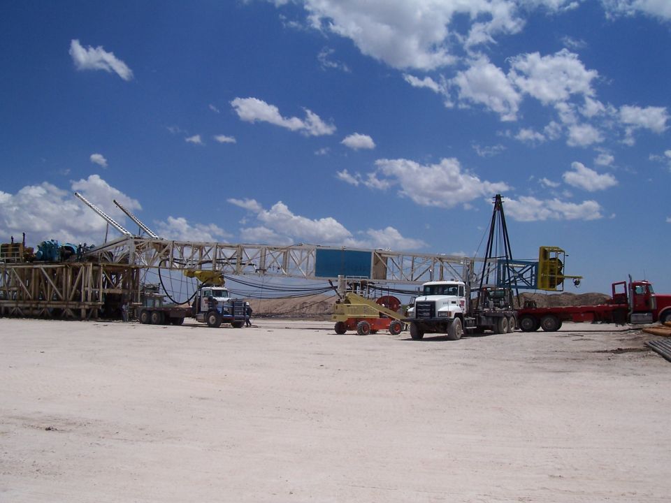 Several trucks and heavy equipment are on a dusty lot under a blue sky with clouds. A large structure that looks like part of an oil rig is in the background