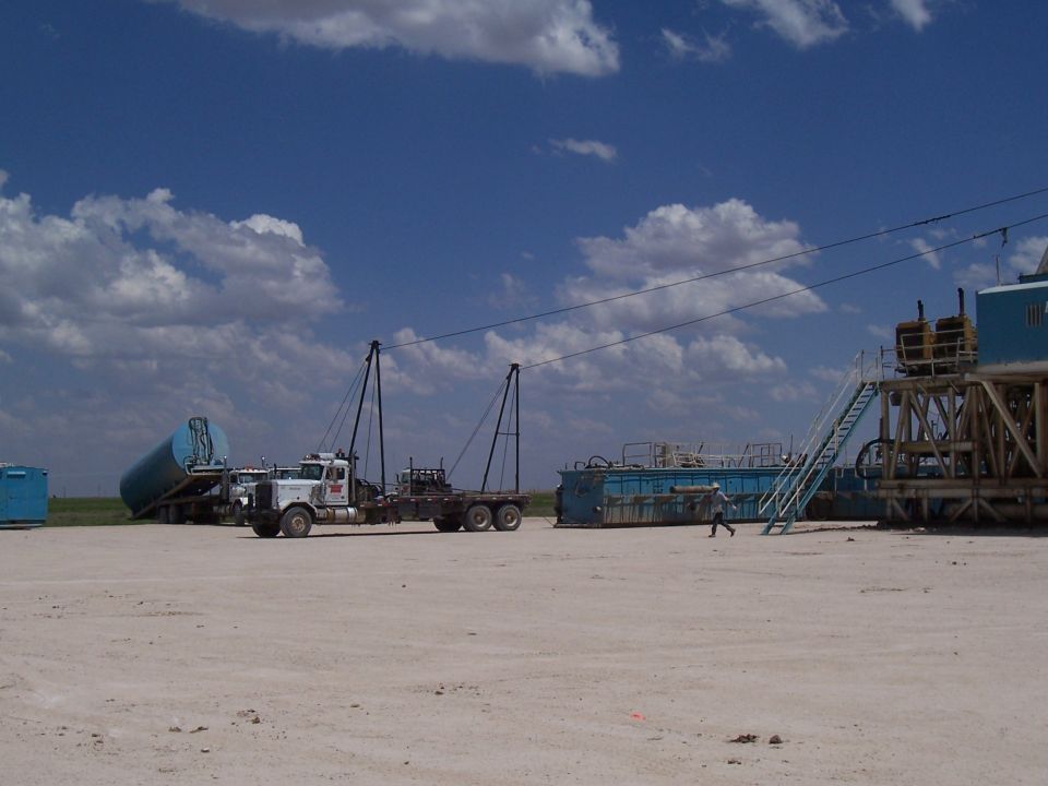 White semi-truck with a flatbed trailer carrying a blue cylindrical tank, parked on a dirt lot next to industrial equipment