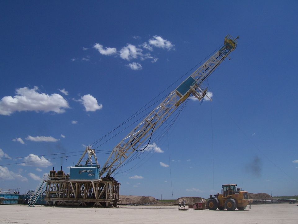 oil drilling rig with 'BIG DOG 400' label, yellow loader in foreground, under blue sky with clouds