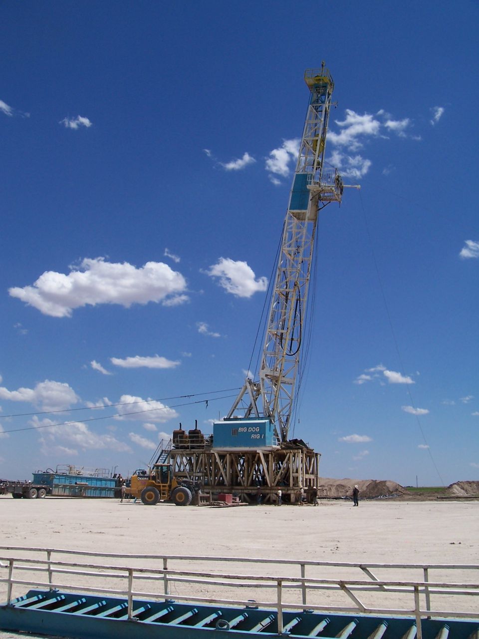 Oil drilling rig with a forklift nearby under a blue sky with clouds