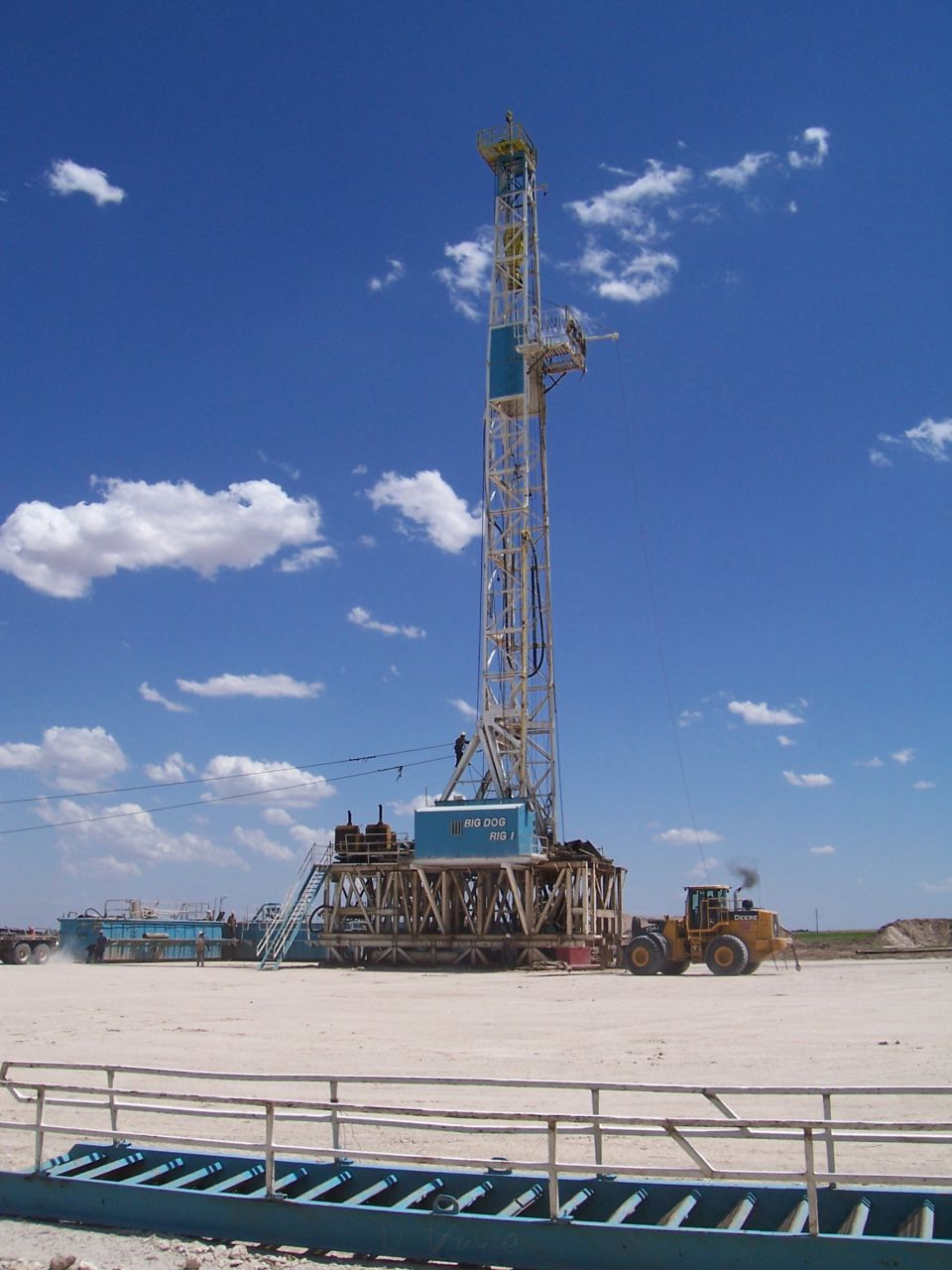 Oil drilling rig under a blue sky with clouds. A yellow front loader is nearby