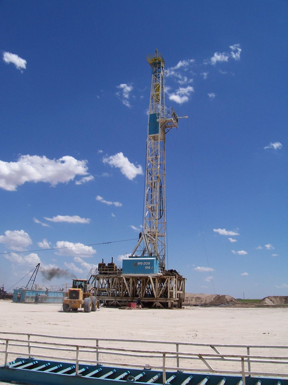 Blue drilling rig 'Big Dog Rig 1' with yellow loader on sandy oilfield