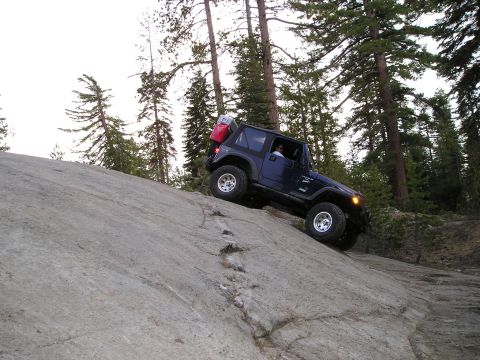 1989 Ford Bronco - Jeep Wrangler navigates a steep, rocky incline in a wooded area.