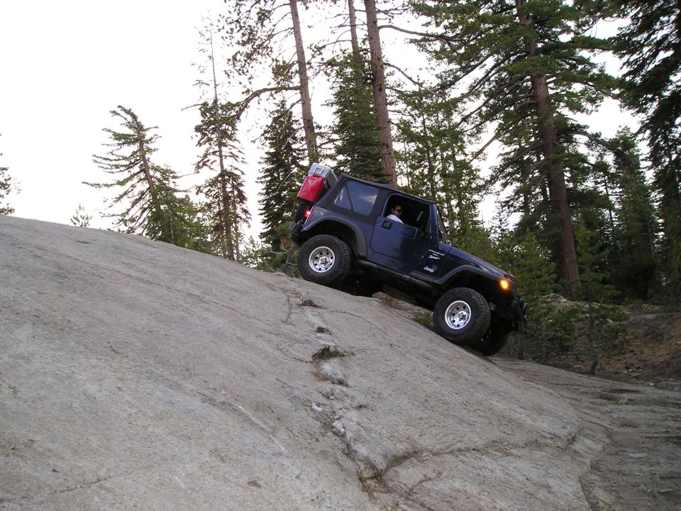 Dark blue Jeep Wrangler climbing a large rock face, spare tire and red gas can mounted on rear