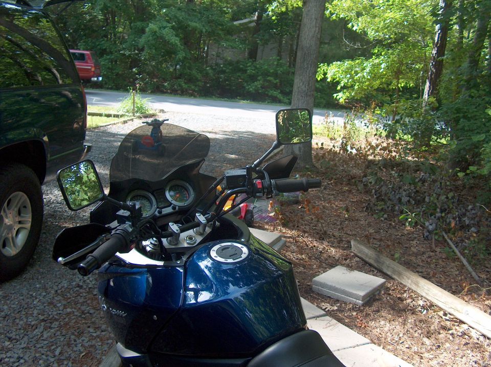 Blue motorcycle's handlebars, mirrors, and fuel tank are visible. A dark SUV is parked to the left