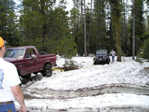 1989 Ford Bronco - Two vehicles are stuck in the snow in a wooded area, with one attempting to tow the other out.