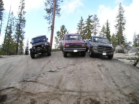 1989 Ford Bronco - Three off-road vehicles are parked on a rocky incline in a forest setting.