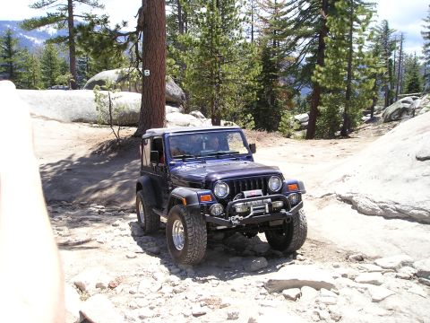 1989 Ford Bronco - A Jeep Wrangler TJ navigates a , mountainous terrain with pine trees and large boulders.