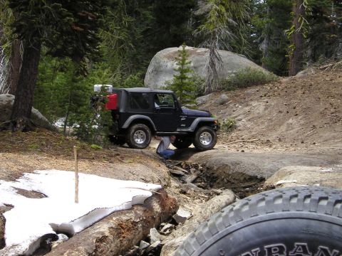 1989 Ford Bronco - A black Jeep Wrangler navigates a rough trail in a wooded area, with a person working on its underside.