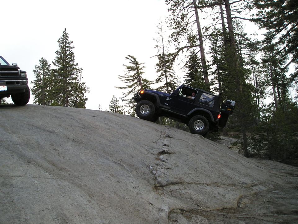 Dark blue Jeep Wrangler climbing a large rock face, with a dark pickup truck visible on the left