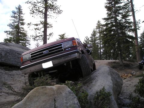 1989 Ford Bronco - A maroon Ford Bronco is shown off-roading over large rocks in a forest.