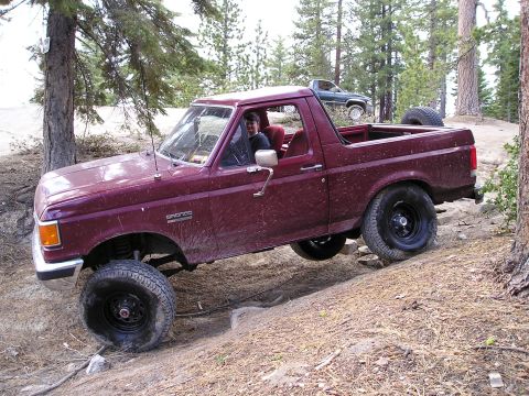 1989 Ford Bronco - Maroon full-size Ford Bronco with the rear top removed, photographed from the driver's side while flexing over a dirt rise in a pine forest…