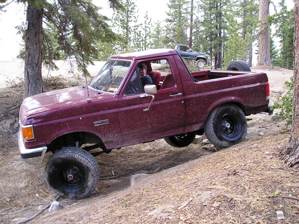 Maroon topless Ford Bronco flexing on a dirt rise in a pine forest, side view
