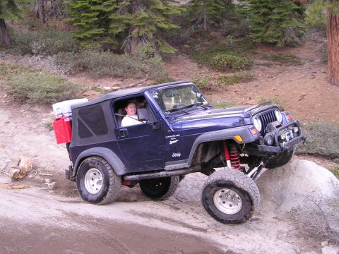 1989 Ford Bronco - A blue Jeep Wrangler navigates a rocky trail in a wooded area, equipped with extra fuel cans and storage.