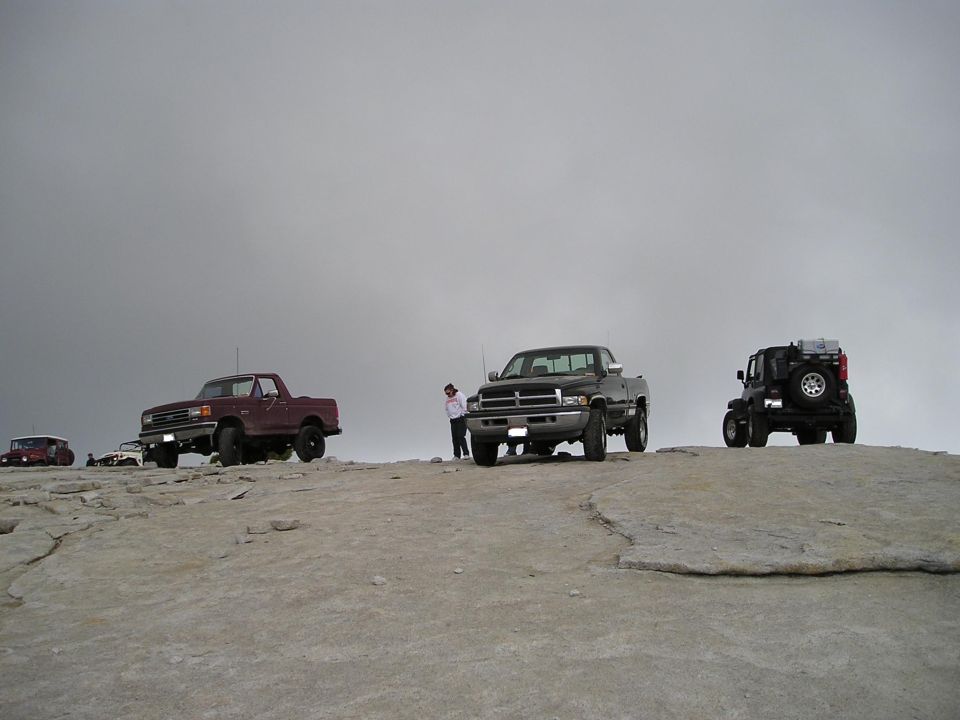 Several trucks and a Jeep parked on a rocky, elevated area under a cloudy sky