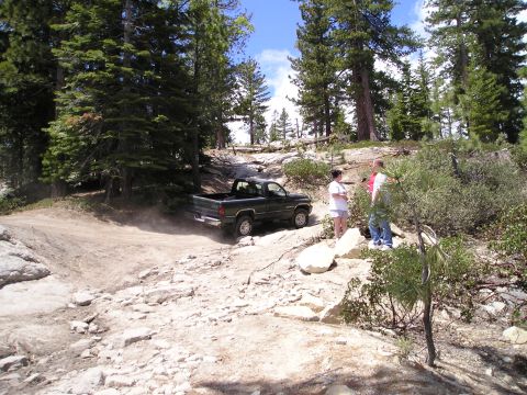 1989 Ford Bronco - A dark green pickup truck navigates a dusty, off-road trail surrounded by trees and rocks.