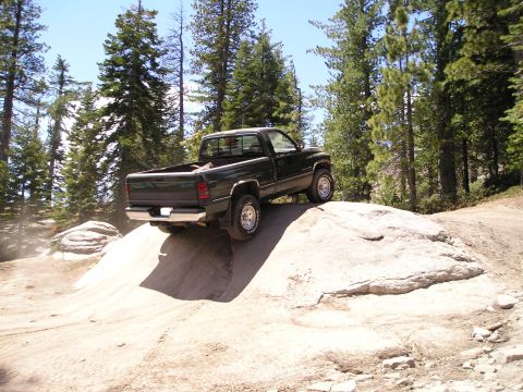 1989 Ford Bronco - A dark green pickup truck is shown driving on a challenging rocky terrain in a wooded area.