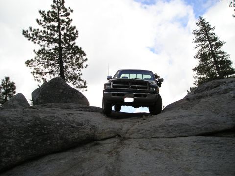 1989 Ford Bronco - Dramatic low front view, shot from below, of a dark Dodge Ram pickup silhouetted on a granite slab against a cloudy sky, with pines flankin…