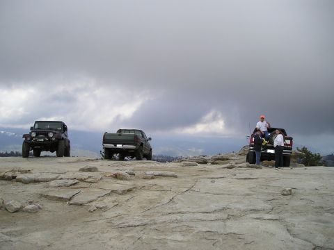 1989 Ford Bronco - Three off-road vehicles are parked on a rocky surface with a cloudy sky overhead.