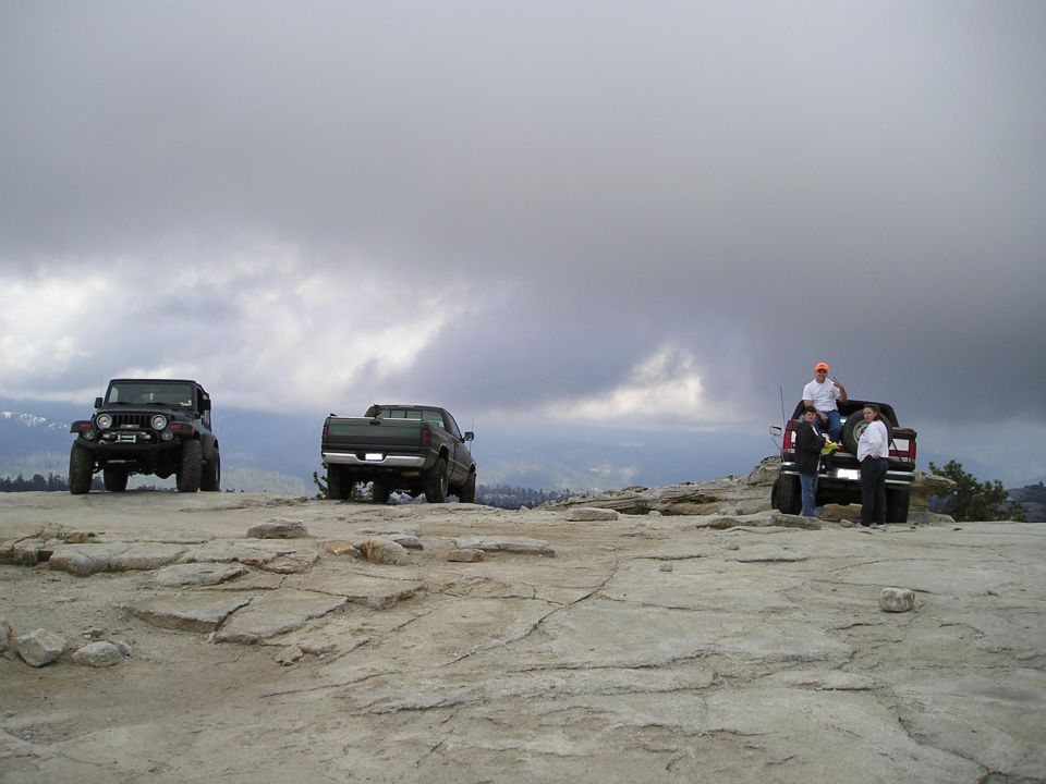Jeep Wrangler and two pickup trucks on a rocky plateau under a cloudy sky