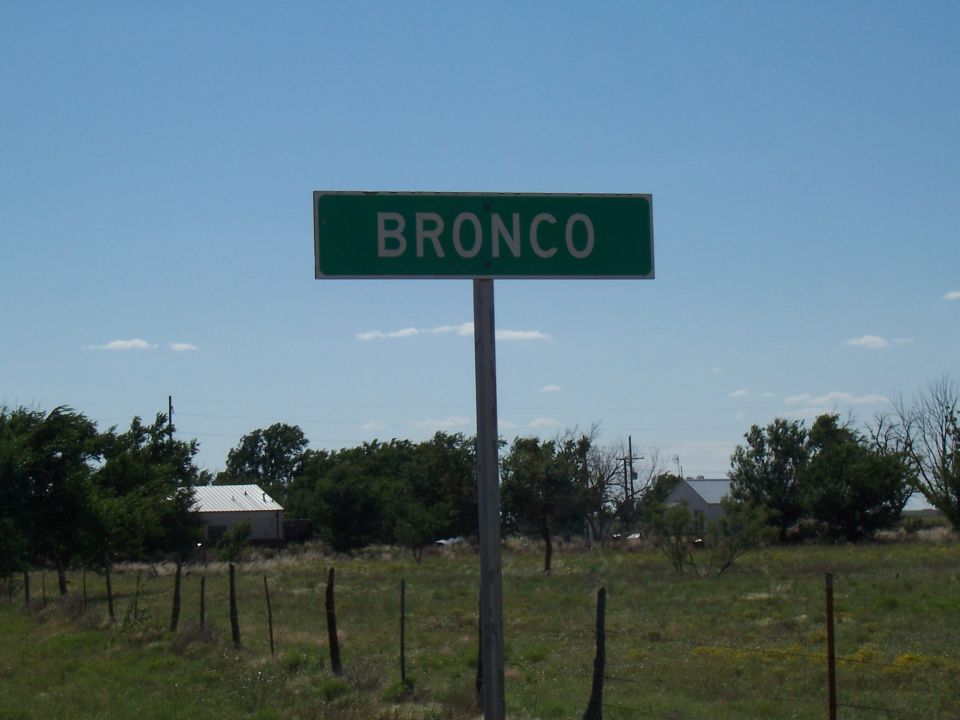 Green street sign that says BRONCO. Field and some buildings in background