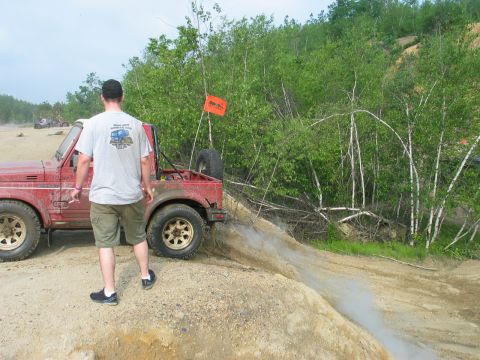 1995 Ford Bronco - Mud-splattered red Suzuki Samurai-style soft-top 4x4 parked at the crest of a sand-and-gravel hill at an off-road park, with a man in a gra…