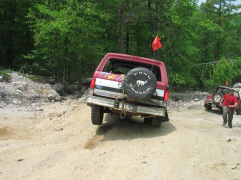 1995 Ford Bronco - Rear view of a maroon full-size Ford Bronco on a rocky dirt trail, with a rear-mounted spare wearing a 'Ford Bronco' bucking-horse cover, a…