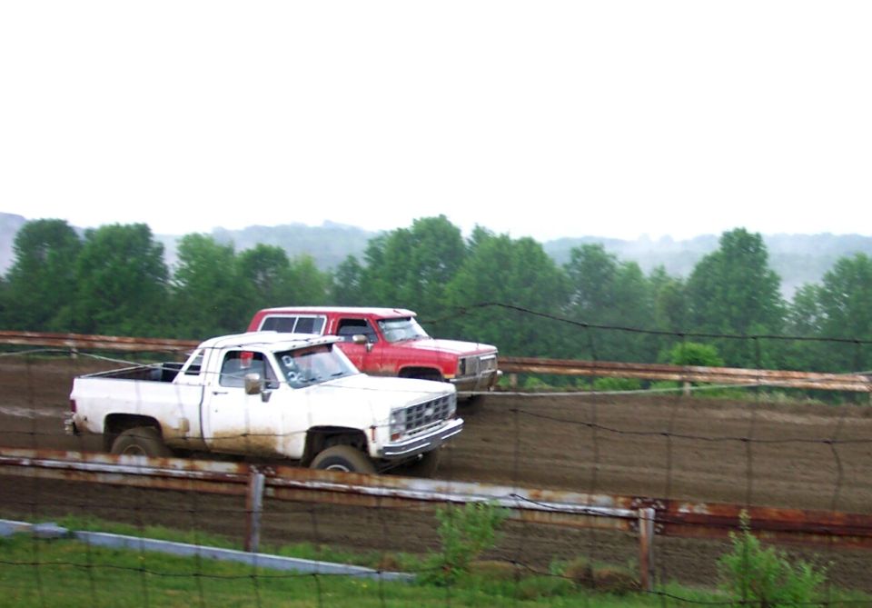 White and red square-body Chevy pickups racing side by side on a muddy dirt track behind fence