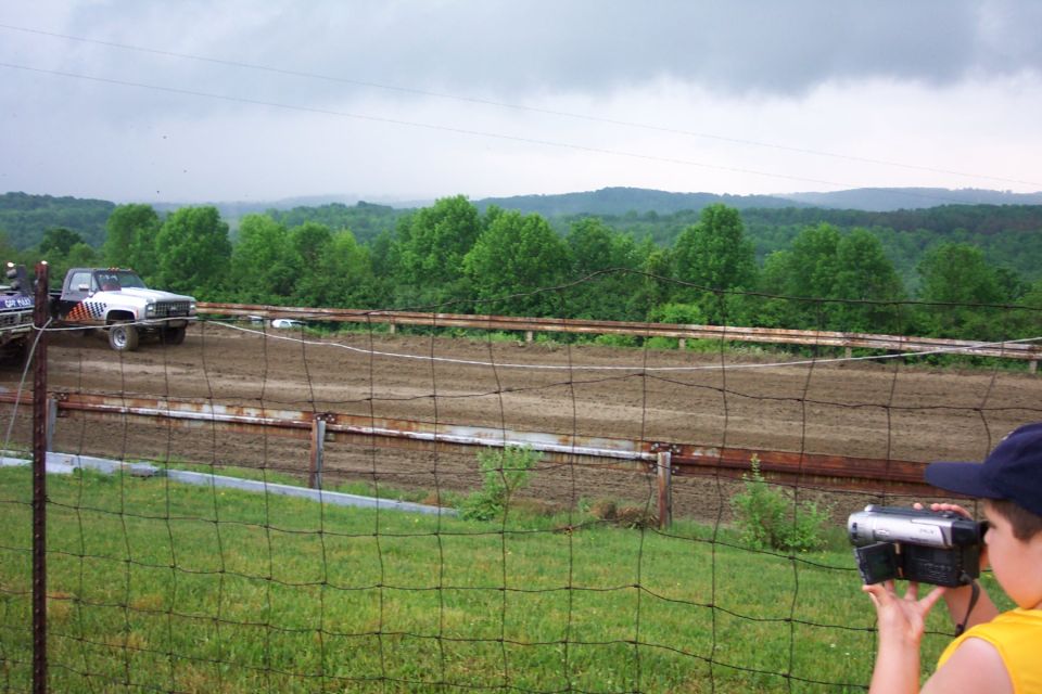 Lifted older pickup truck on a muddy dirt arena track viewed from behind a wire fence with hills beyond