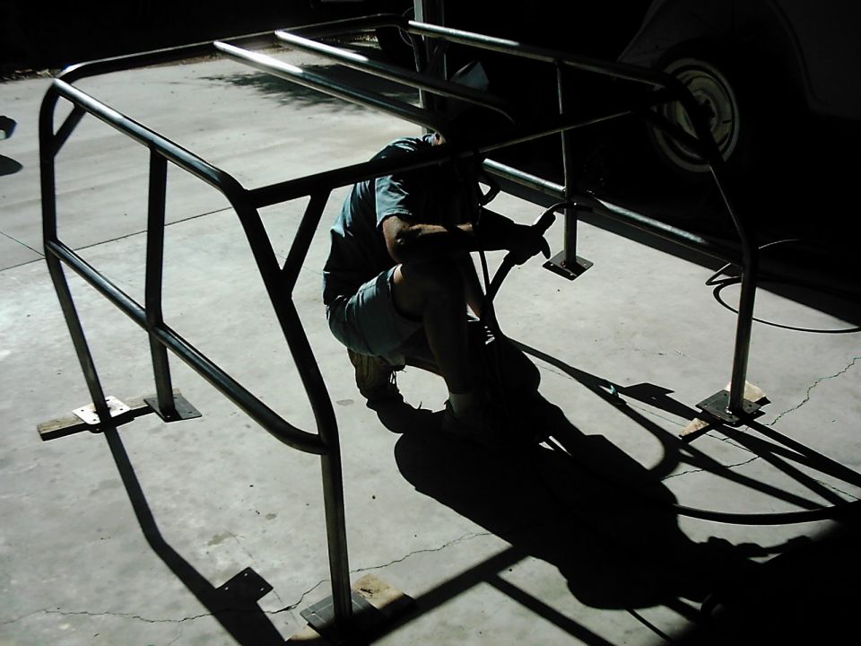 Roll cage being installed on concrete floor, with a person working on it. Part of a vehicle is visible in the background