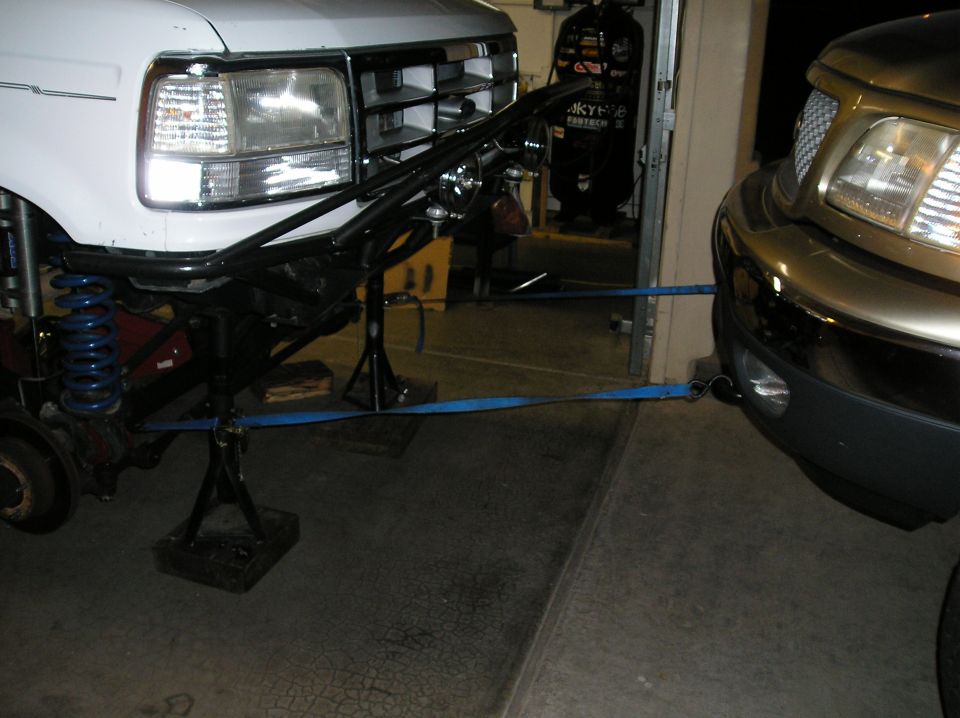 White Ford on jack stands with tube bumper and blue coil springs, strapped to a gold F-150 in a garage