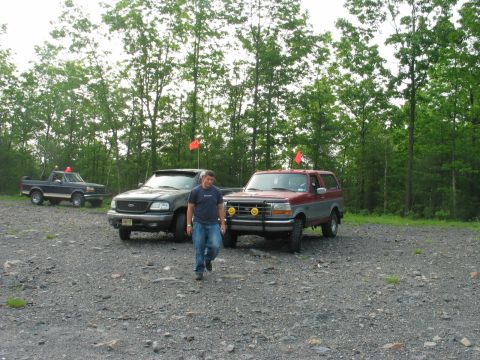 1995 Ford Bronco - Three trucks parked on a gravel staging area at the edge of the woods: a two-tone maroon and silver full-size Ford Bronco with a brush guar…