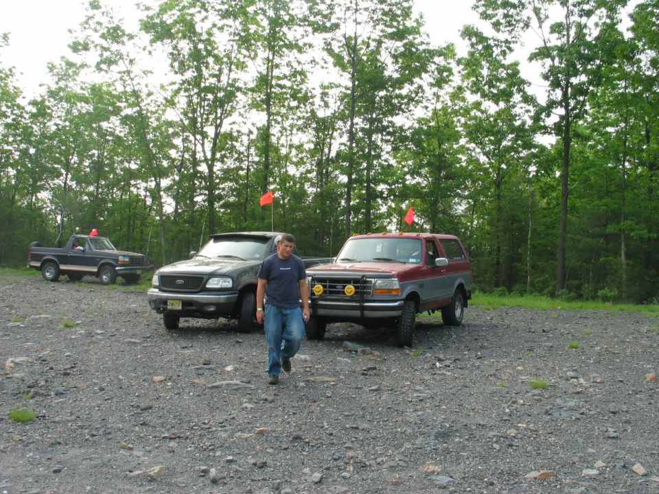 Maroon Ford Bronco with brush guard and trail flag parked with other trucks on gravel