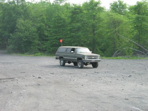 1995 Ford Bronco - Olive-drab square-body Chevrolet Suburban 4x4 parked alone on a gray gravel lot at the edge of green woods, flying a tall orange whip flag…